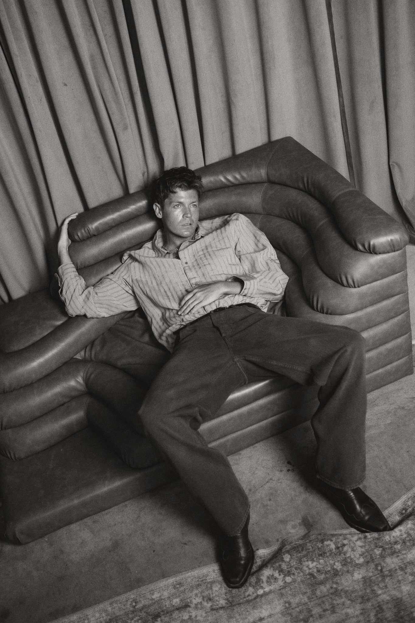 Jamie Miller reclining on a curved leather sofa, black and white